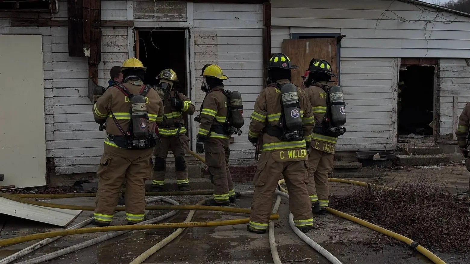 Firefighters in turnout gear gathered at a doorway with hose lines on the ground.