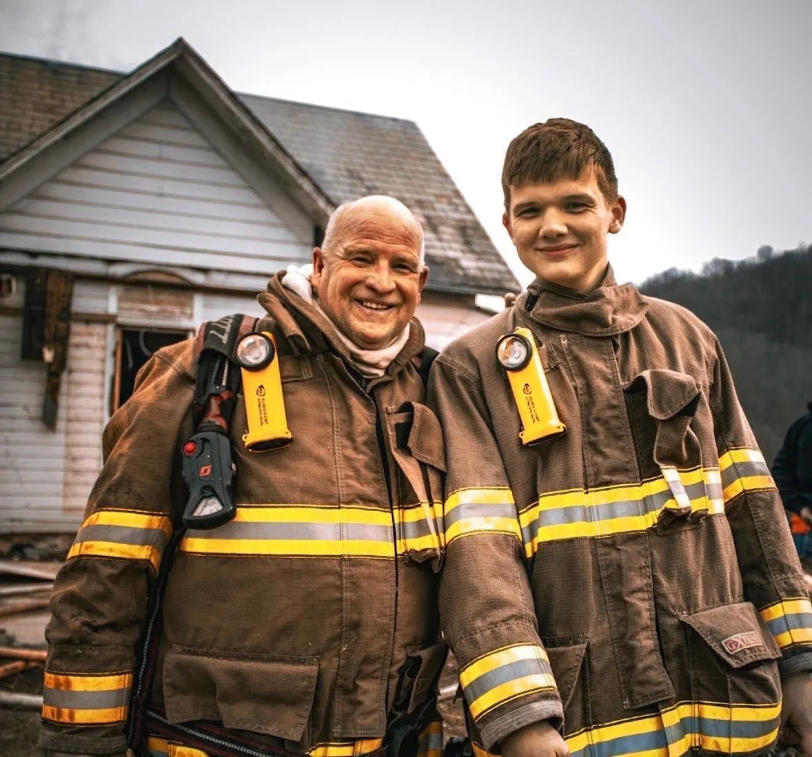 Two firefighters in turnout gear standing together outdoors.