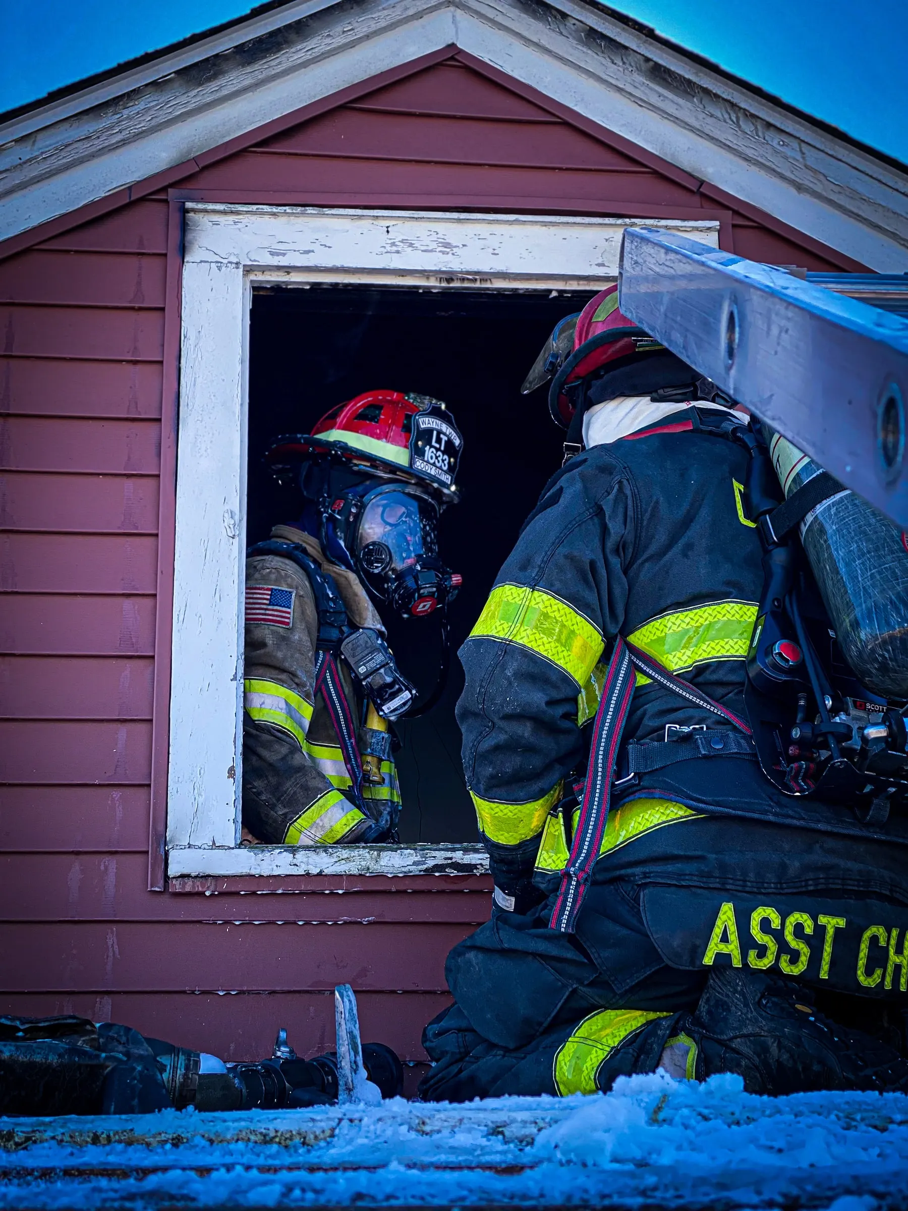 Firefighters in turnout gear entering a window during a structure-fire response.