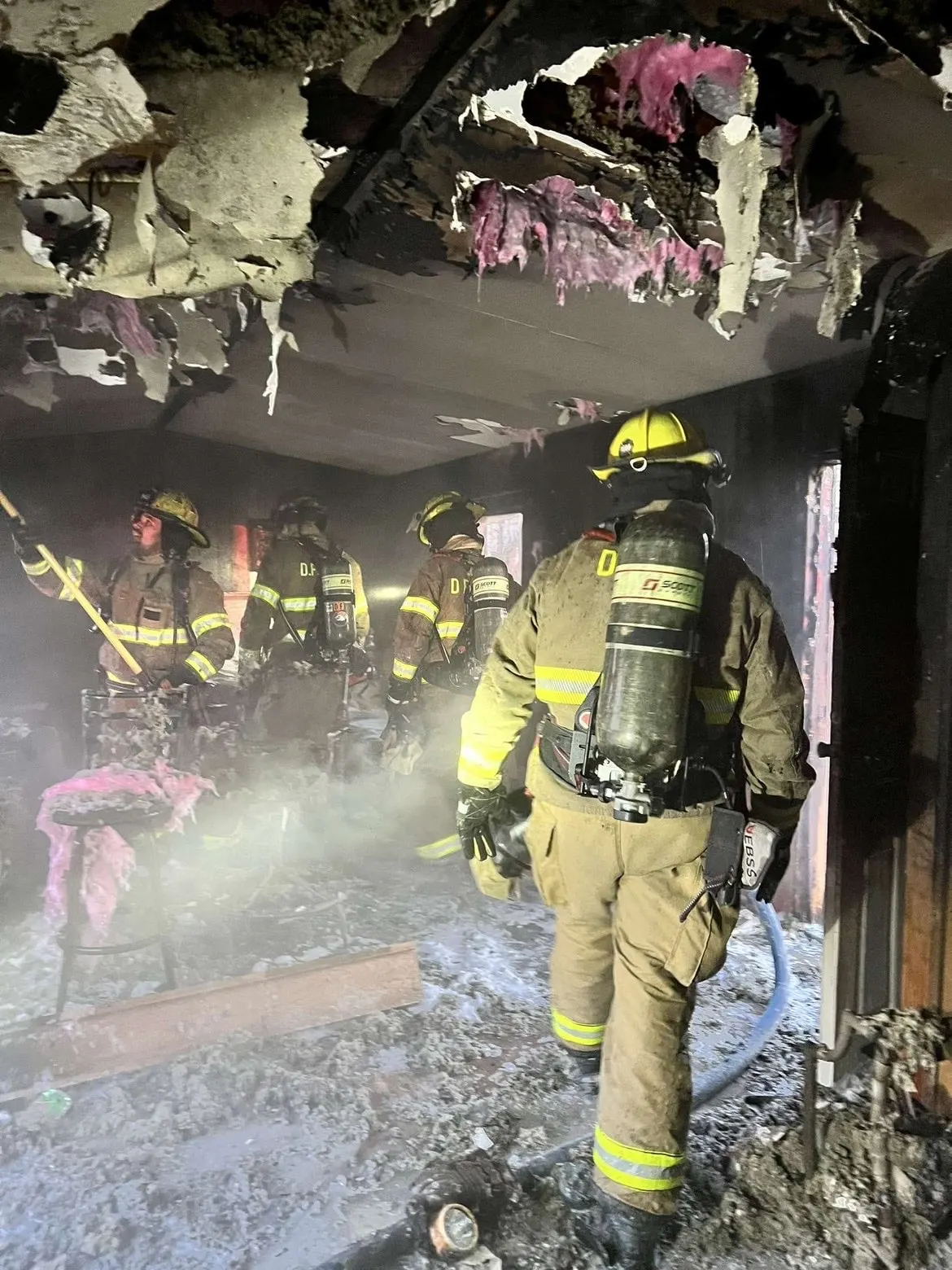 Firefighters in turnout gear working inside a smoke-damaged room.