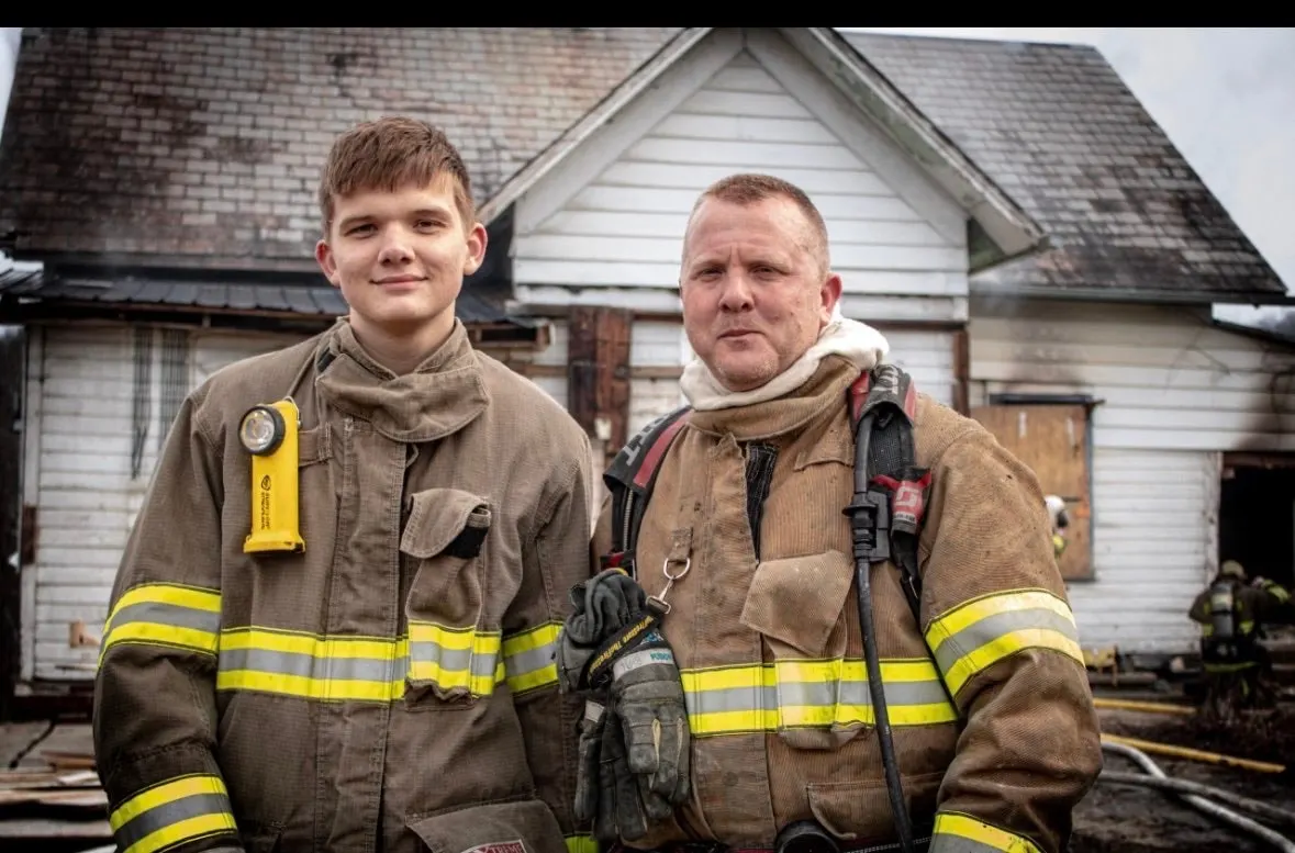Two firefighters in turnout gear standing in front of a house.