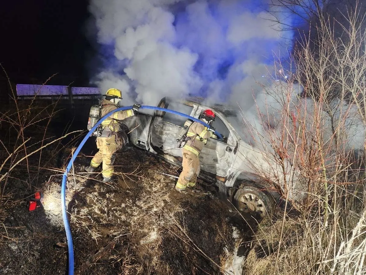 Firefighters using a hose near a burned vehicle at night.